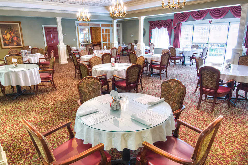 A spacious dining room with multiple round tables covered with white tablecloths and glass tops. Each table is set with menus, napkins, and small condiment holders. The room features patterned carpet, upholstered chairs with wooden arms, chandeliers hanging from the ceiling, large windows with red curtains, and decorative columns.