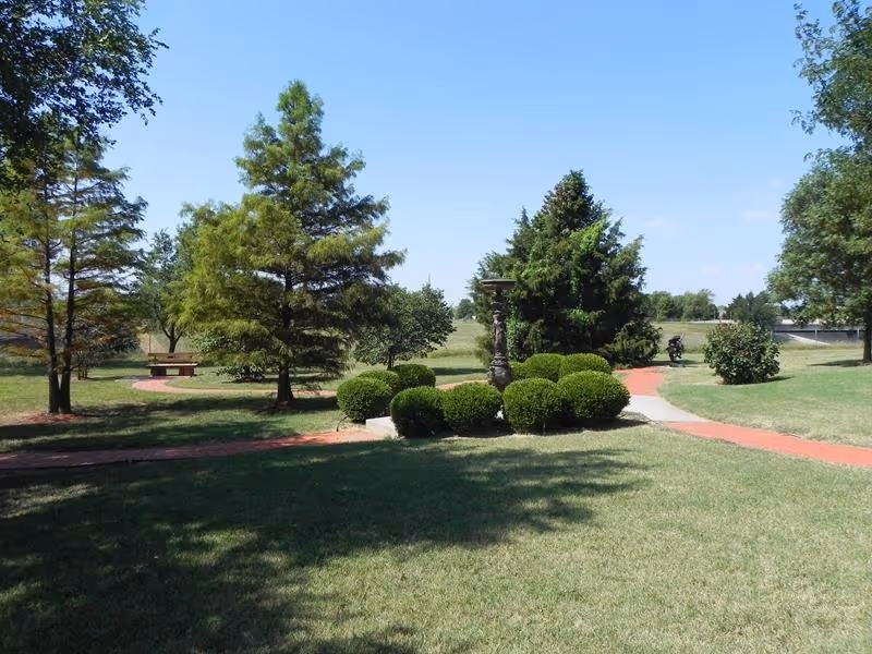 A landscaped outdoor area with green grass, trimmed bushes, several trees, and a paved walkway winding through the space under a clear blue sky.