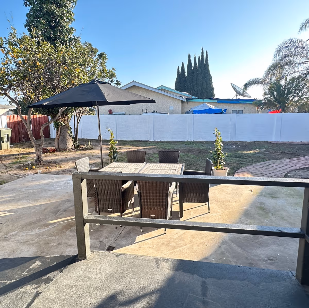 Outdoor patio area with a rectangular table and six wicker chairs under a large black umbrella. The patio is surrounded by a concrete surface and a small garden area with trees, plants, and a white fence in the background. A house and tall cypress trees are visible beyond the fence.