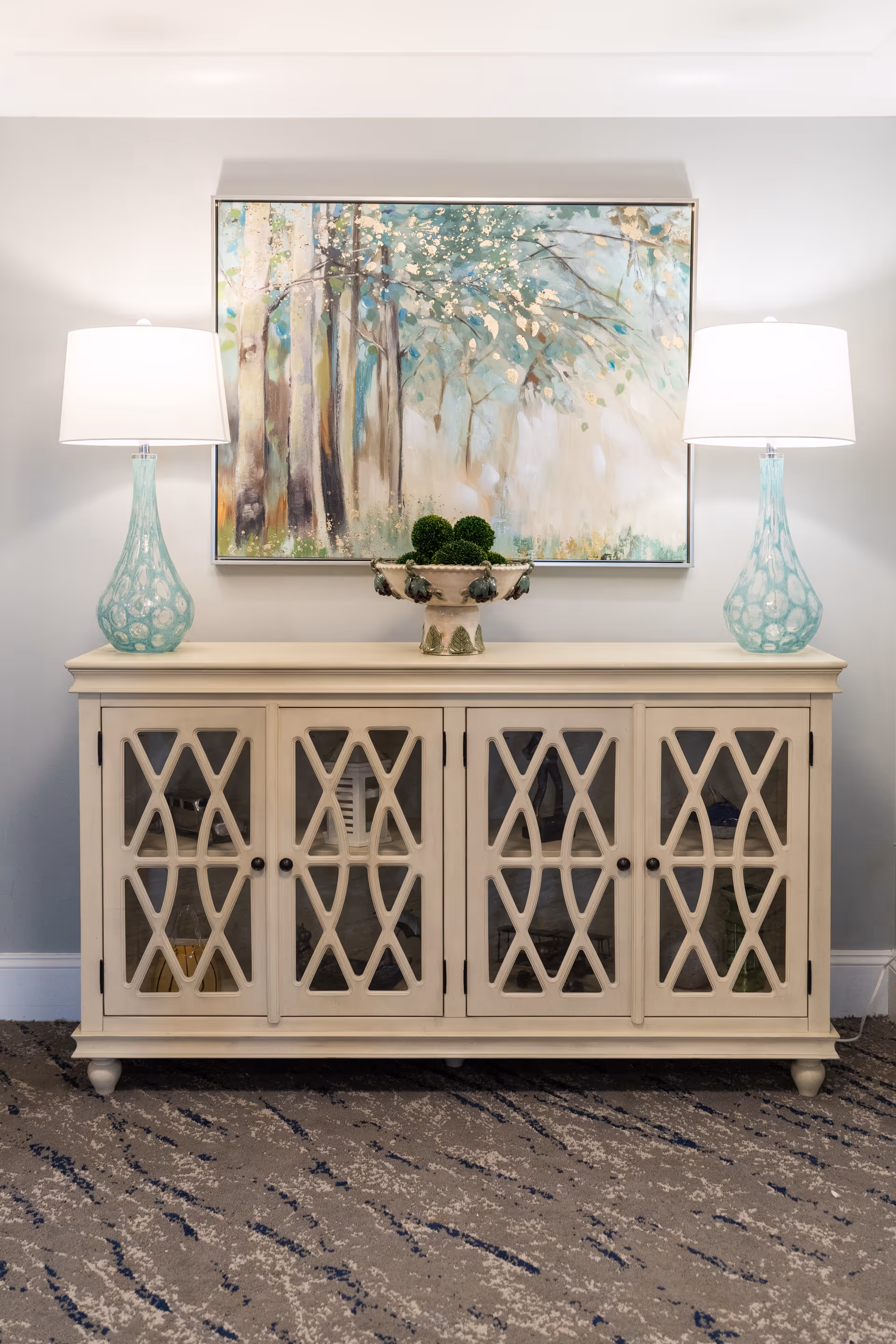 A beige wooden cabinet with glass doors featuring a crisscross design, topped with a decorative bowl containing green moss balls. On either side of the bowl are two matching turquoise glass lamps with white lampshades. Above the cabinet hangs a large painting of a forest scene with trees and green foliage. The floor is carpeted with a patterned design in shades of gray and blue.