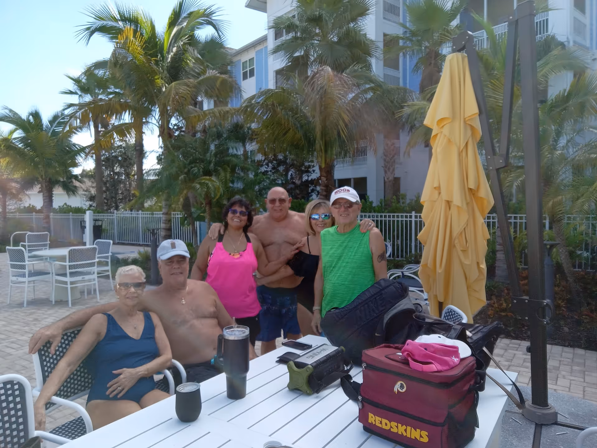 A group of six adults, some in swimwear, gathered around a white outdoor table near a pool area with palm trees and a multi-story building in the background. The table has various personal items including a Redskins cooler bag, hats, and drink containers. The setting appears sunny and tropical.