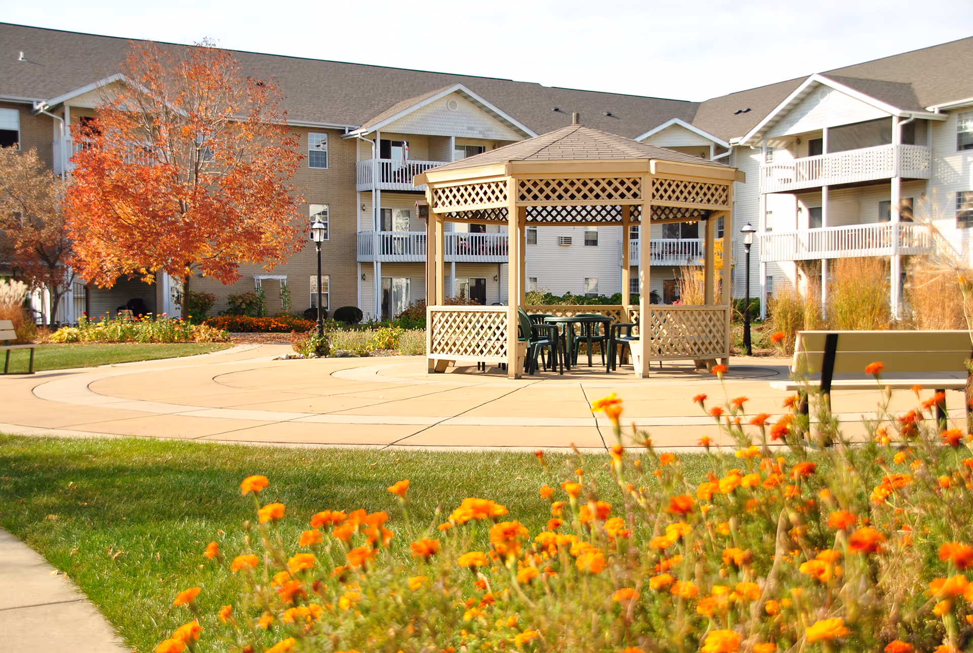Outdoor garden area at Rosewood Villas senior housing community featuring a wooden gazebo with chairs inside, surrounded by green grass, orange flowers, benches, and trees with autumn foliage. Apartment buildings with balconies are visible in the background.