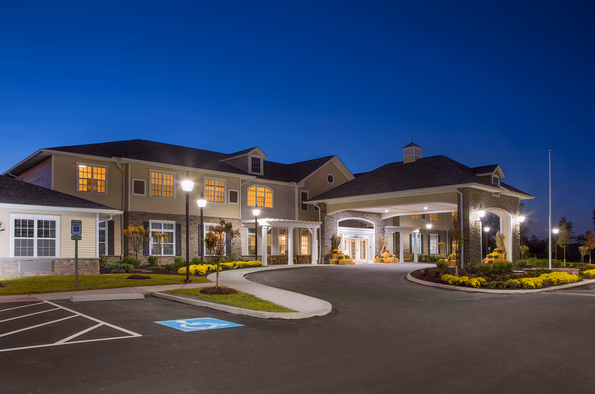 Exterior view of Maumee Pointe Assisted Living and Memory Care facility at dusk, showing a well-lit entrance with a covered drop-off area, landscaped flower beds, and a parking lot with a handicap parking space.