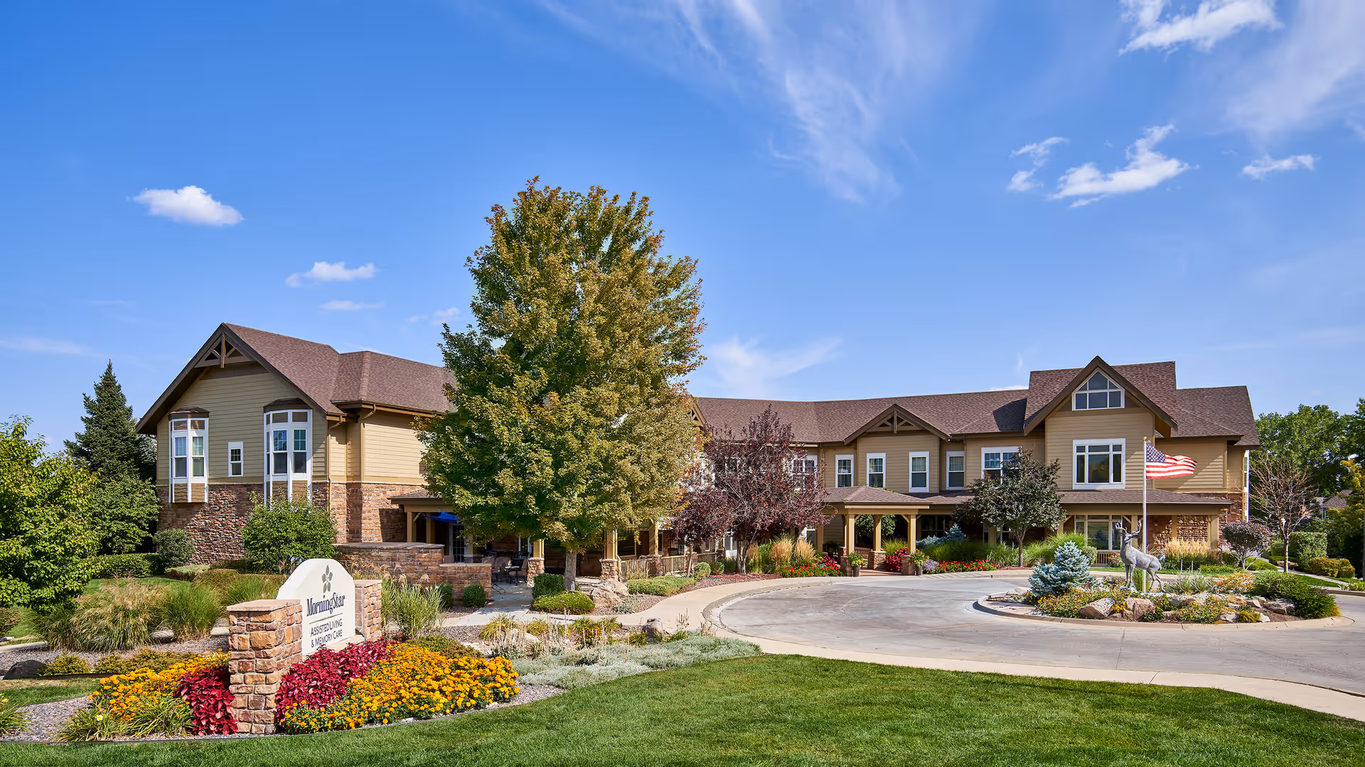 Exterior view of MorningStar Assisted Living & Memory Care of Littleton facility on a sunny day, showing a large two-story building with beige and brown siding, a circular driveway, landscaped gardens with colorful flowers, trees, and an American flag near the entrance.