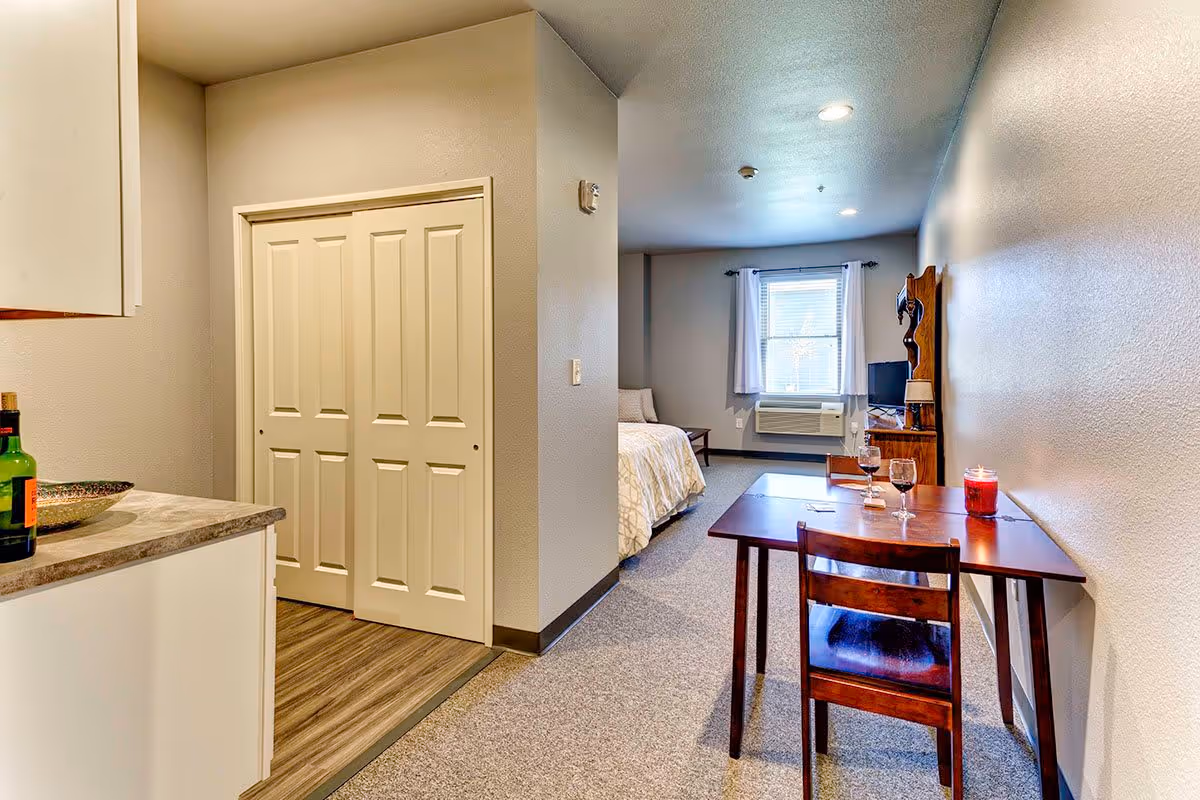 Interior view of a senior living facility apartment showing a small dining table with two chairs, two glasses of wine, and a lit candle. In the background, there is a bed near a window with white curtains, a wooden dresser with a TV, and an air conditioning unit below the window. The floor has a combination of carpet and wood-like flooring, and there is a closet with sliding doors near the kitchen counter.