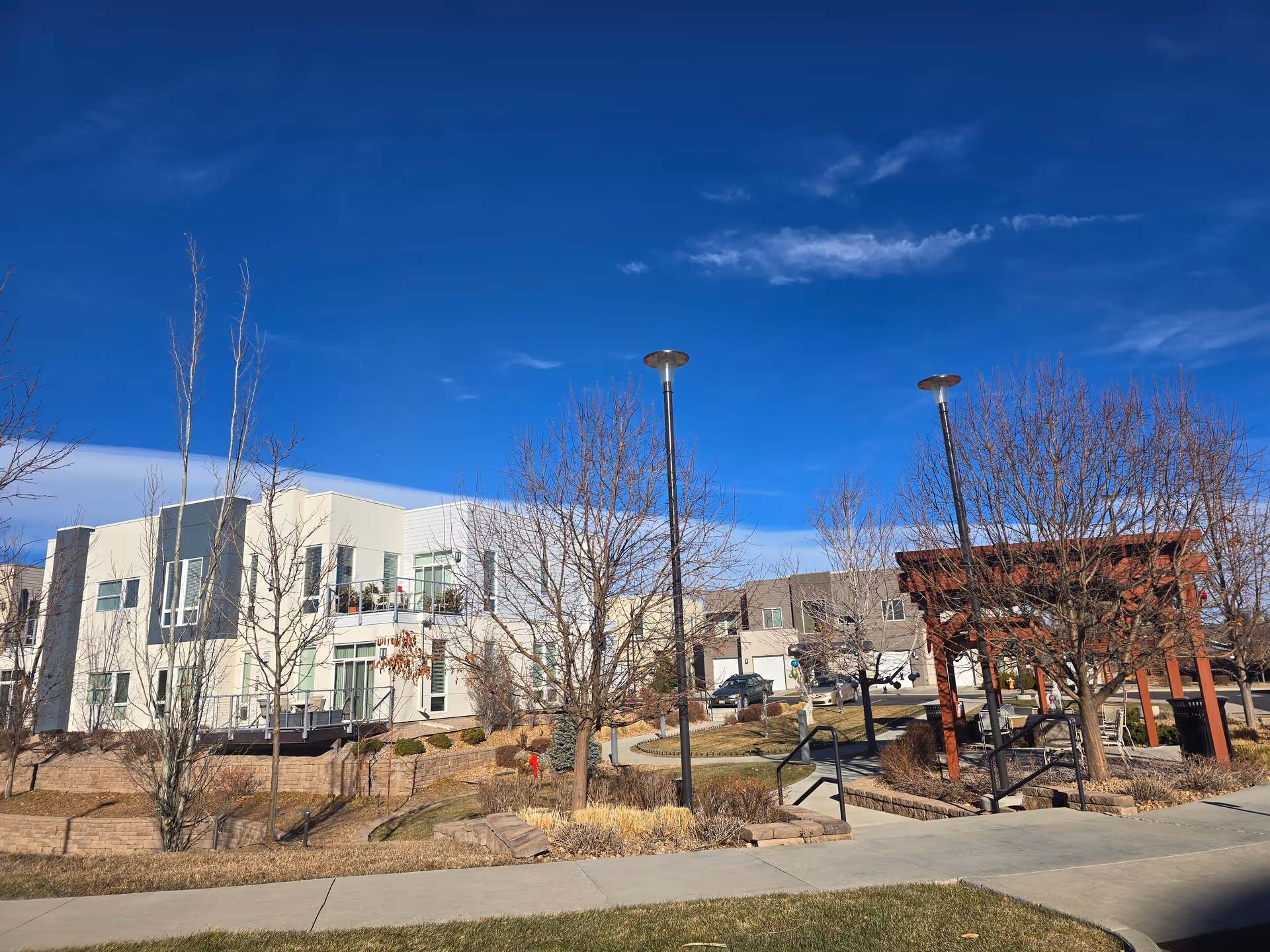 Outdoor view of a senior living facility named Village at Belmar featuring modern two-story buildings, leafless trees, a paved walkway, street lamps, and a wooden pergola structure under a clear blue sky.