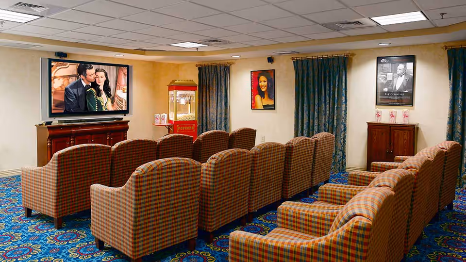 A cozy movie theater room with three rows of plaid upholstered armchairs facing a large flat-screen TV mounted on the wall. The TV is showing a classic movie scene with a man and woman. There is a vintage-style popcorn machine in the corner and framed movie posters on the walls. The room has patterned blue carpet and curtains on the windows.
