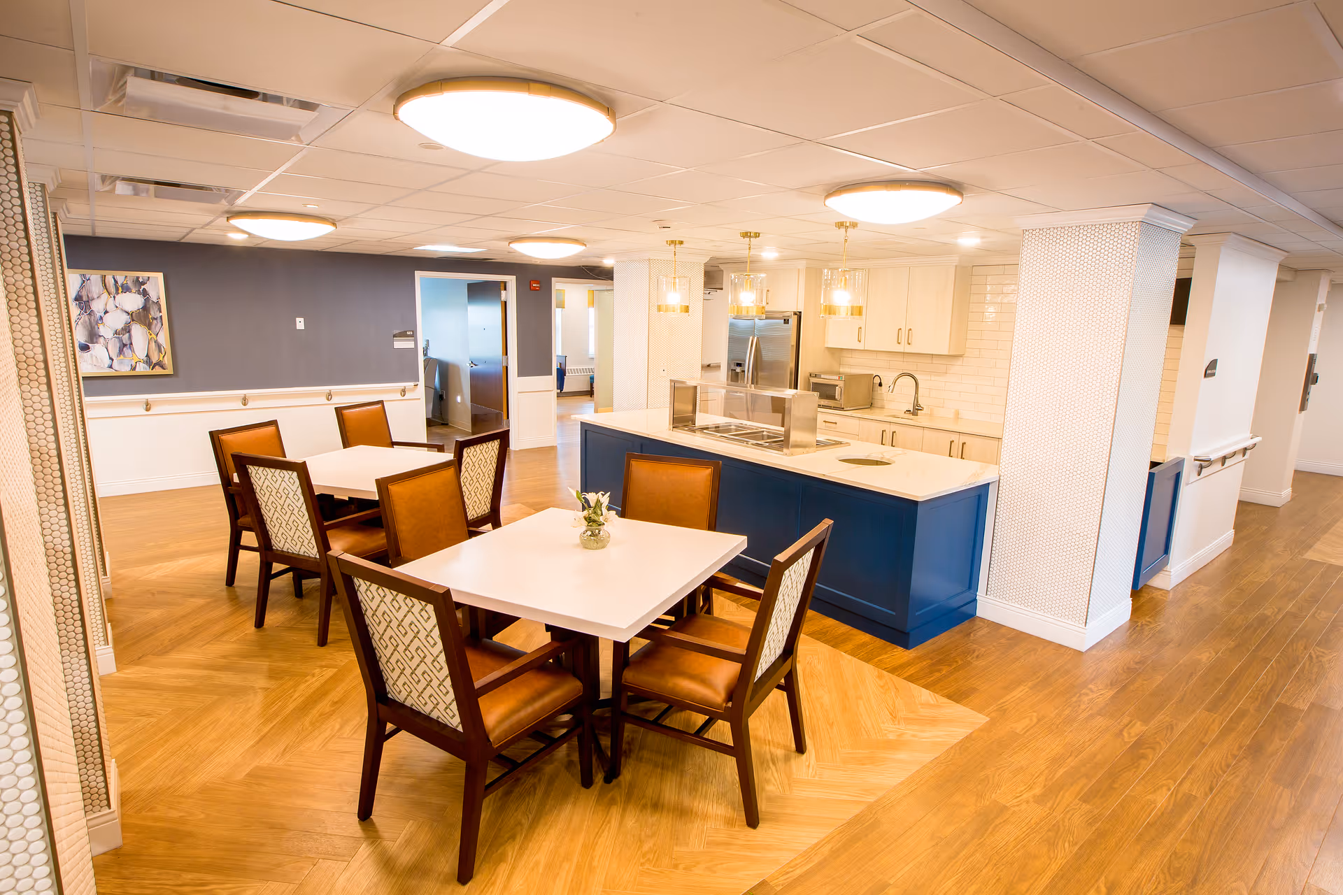 Interior view of a senior living facility dining area with two square tables, each surrounded by four chairs with brown seats and patterned backs. The room features a kitchen island with a blue base and white countertop, equipped with a sink and food service area. The background shows white cabinetry, a stainless steel refrigerator, and pendant lights hanging from the ceiling. The floor is wood with a herringbone pattern near the tables, and the walls are painted in neutral tones with some artwork visible.