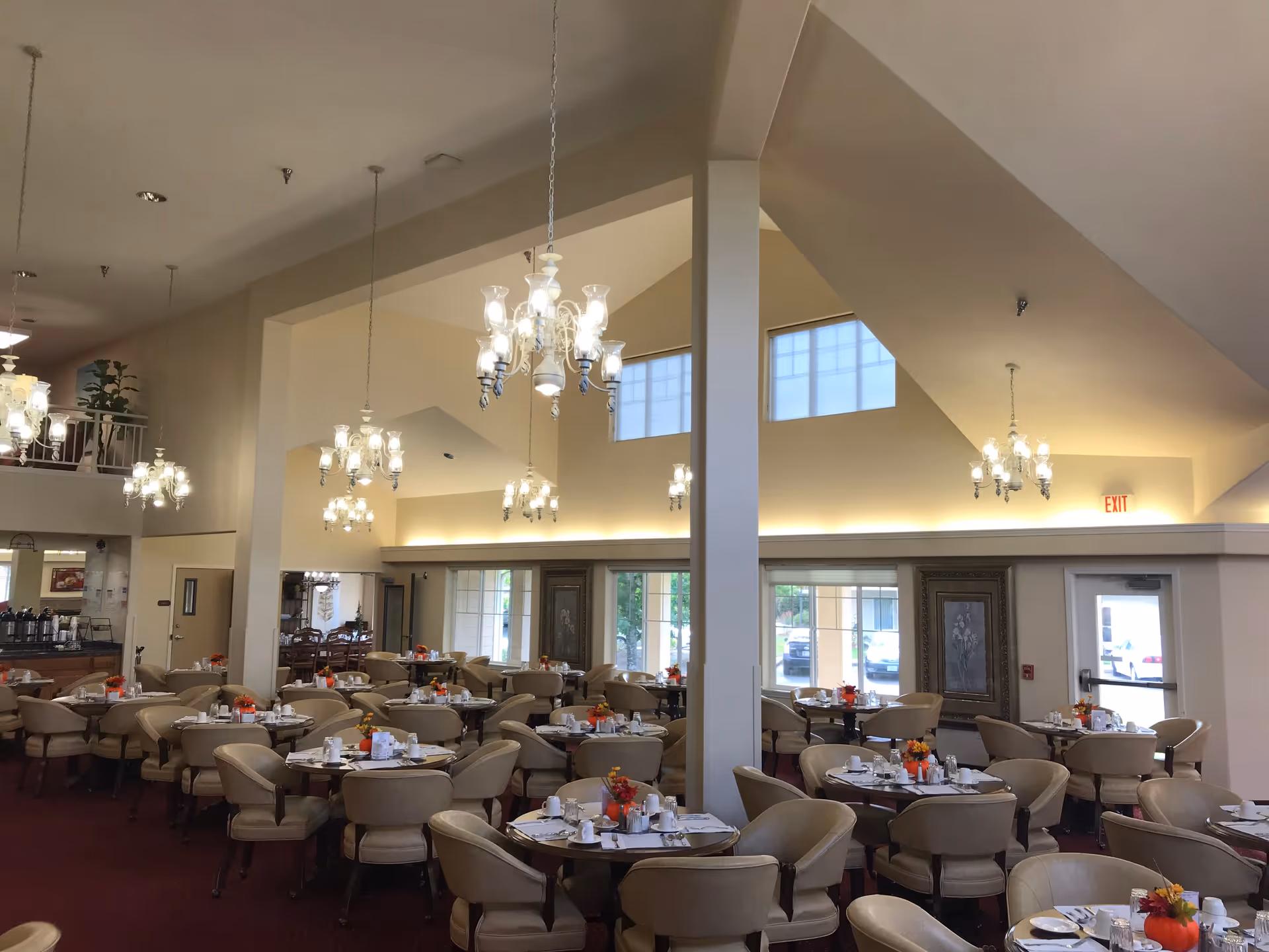 A spacious dining room with multiple round tables set for meals, each table decorated with small orange flower arrangements. The room features high vaulted ceilings with several elegant chandeliers hanging down, large windows letting in natural light, and beige walls. Comfortable beige chairs surround each table, and there is an exit door visible on the right side.