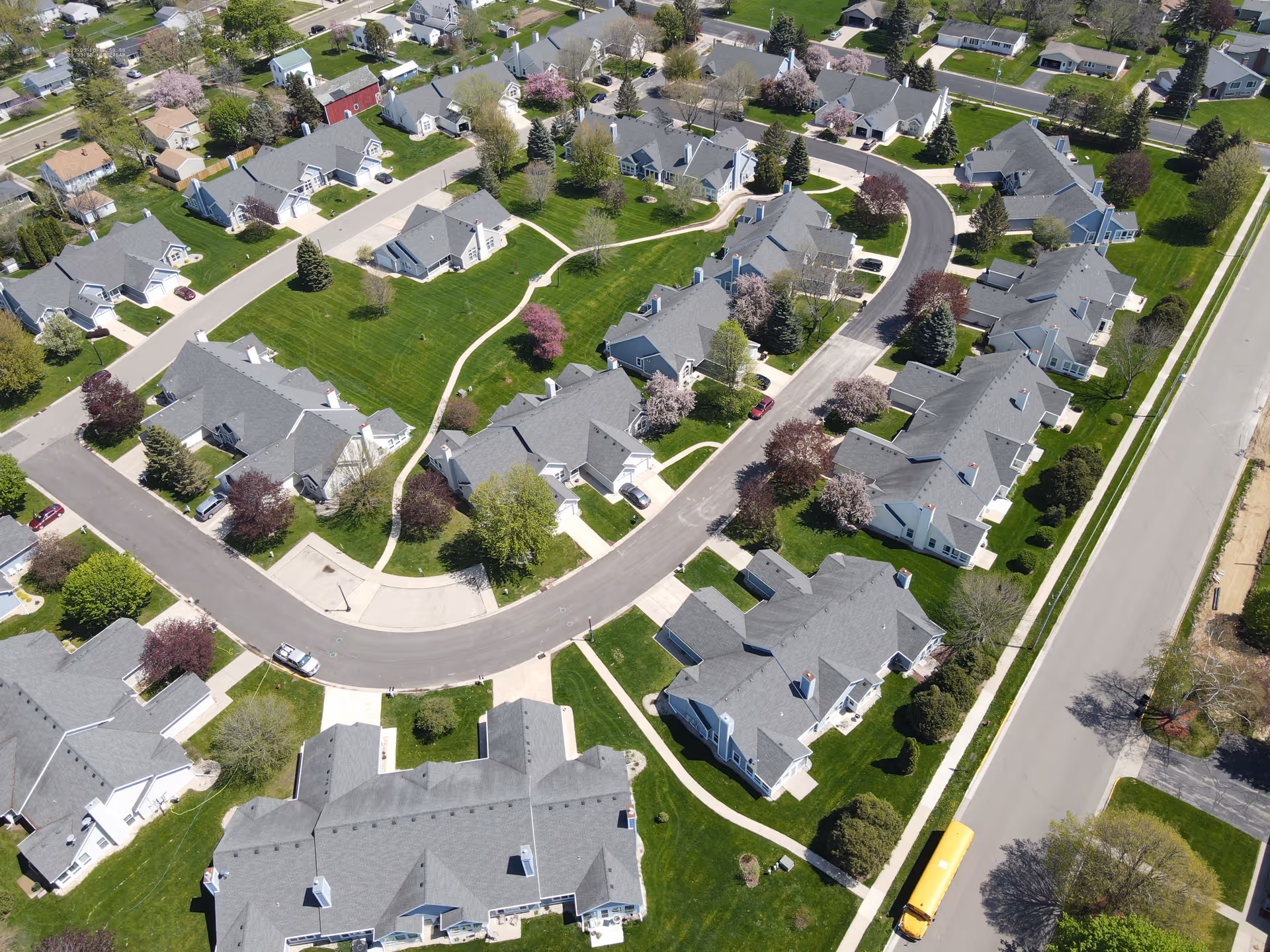 Aerial view of a residential neighborhood with multiple houses, green lawns, trees with spring blossoms, and curved roads. A yellow school bus is visible on the road on the right side of the image.