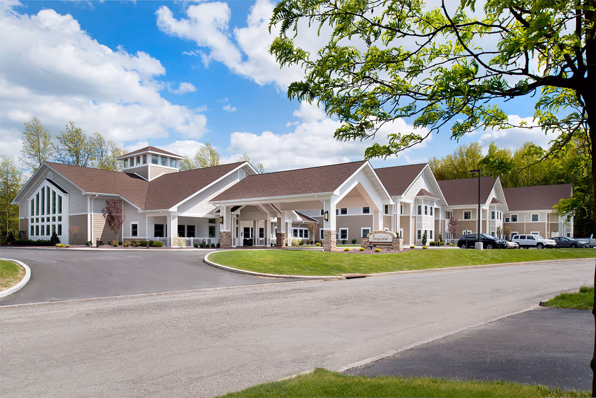 Exterior view of The Inn at Ironwood, a large, modern senior living facility with beige siding and brown roofs, surrounded by green grass and trees under a partly cloudy blue sky.