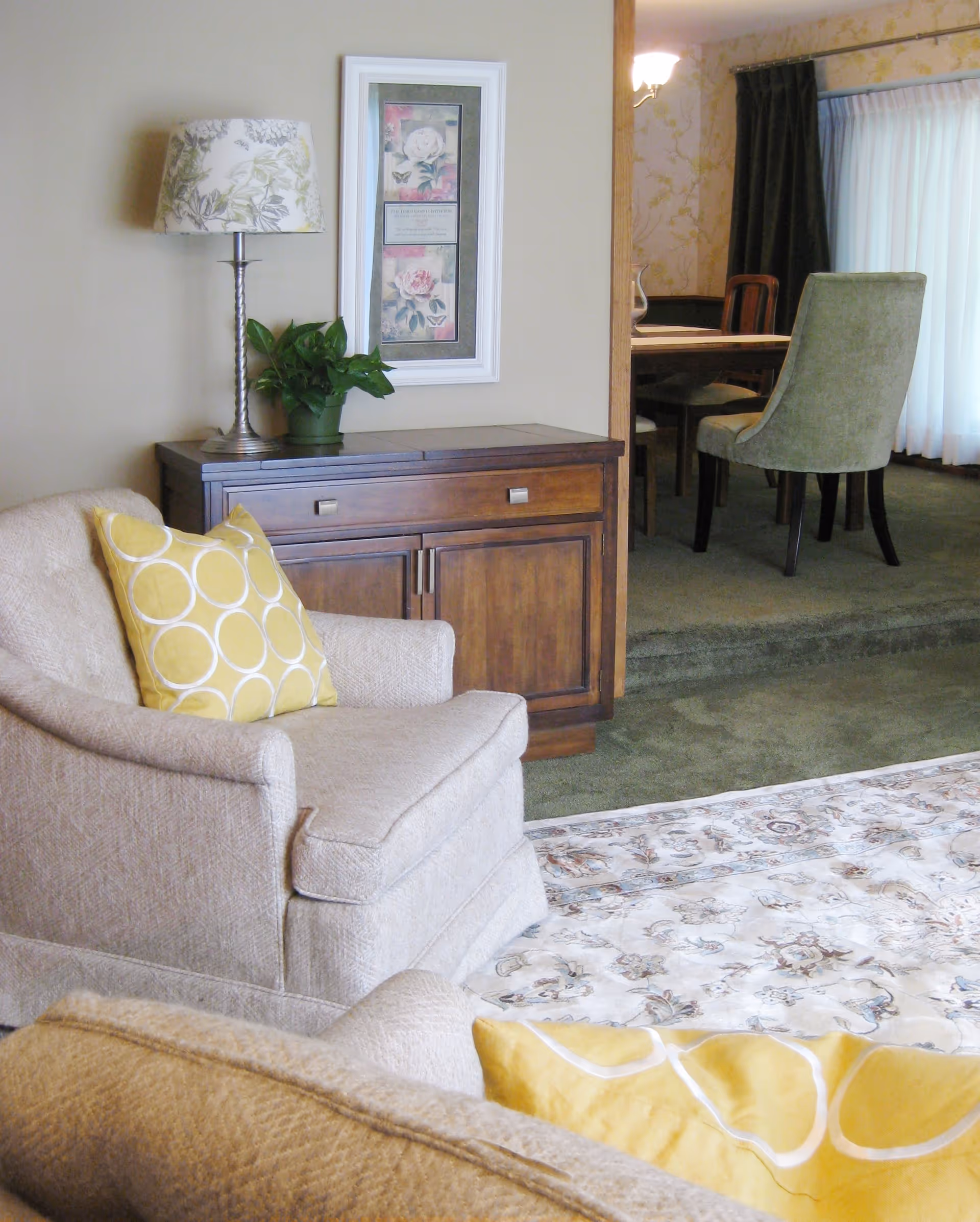 A cozy living room area with a beige armchair featuring a yellow pillow with white circles, a wooden cabinet with a floral lamp and a green potted plant on top, and a framed floral artwork on the wall. In the background, a dining room with a wooden table and green upholstered chairs is visible, along with a window covered by sheer white curtains and dark drapes.