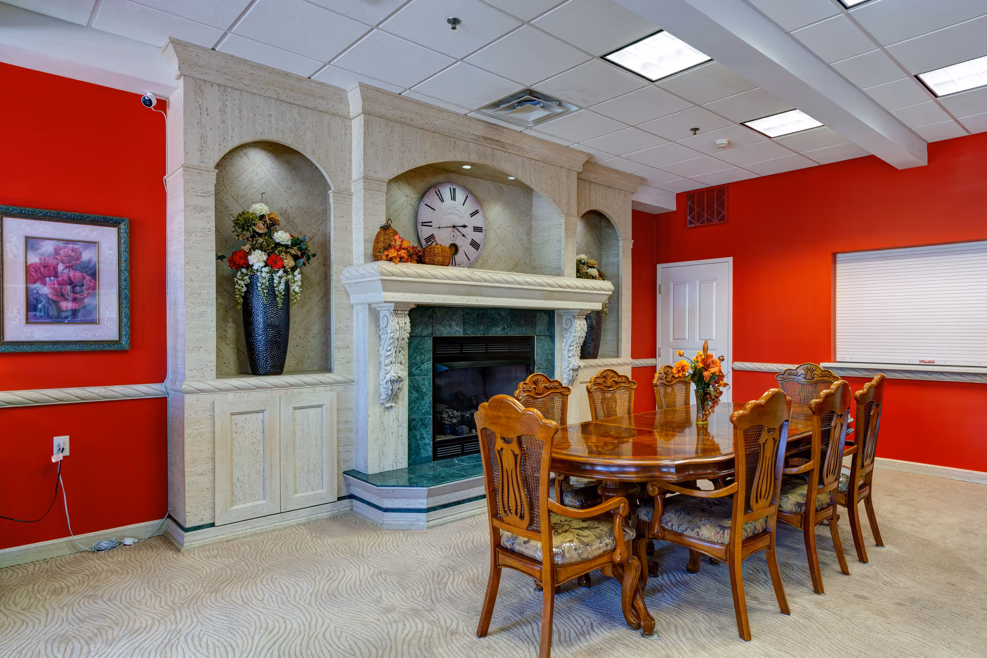 A dining room with a long wooden table surrounded by eight ornate wooden chairs with cushioned seats. The room features a decorative fireplace with a large clock above it and floral arrangements in vases on either side. The walls are painted bright red, and there is a framed floral painting on one wall. The ceiling has recessed lighting and a white drop ceiling.