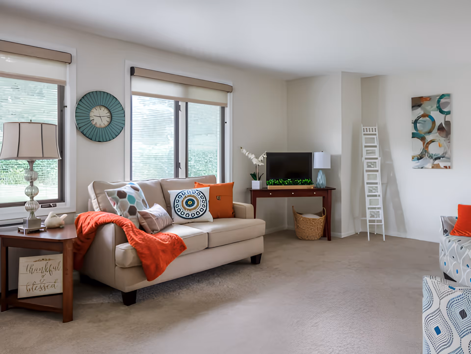 Bright furnished living room with a beige sofa, colorful pillows and throw, side table with lamp, TV on a console, wall art and large windows.
