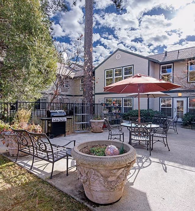 Outdoor patio area at Cascades of Grass Valley featuring metal chairs and tables with a large red umbrella, a barbecue grill, large decorative planters, and a two-story building in the background under a partly cloudy sky.