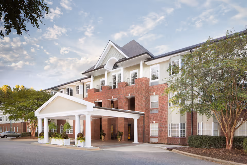 Exterior view of a three-story senior living facility building with a brick and beige facade, a covered entrance with white columns, and surrounding trees under a partly cloudy sky.