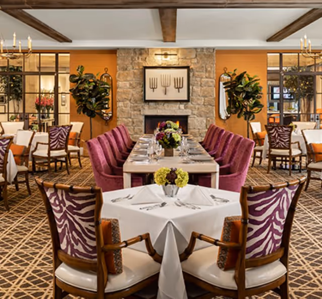 Elegant dining room with a long set table flanked by purple upholstered chairs and a small foreground table, centered around a stone fireplace and decorative plants.