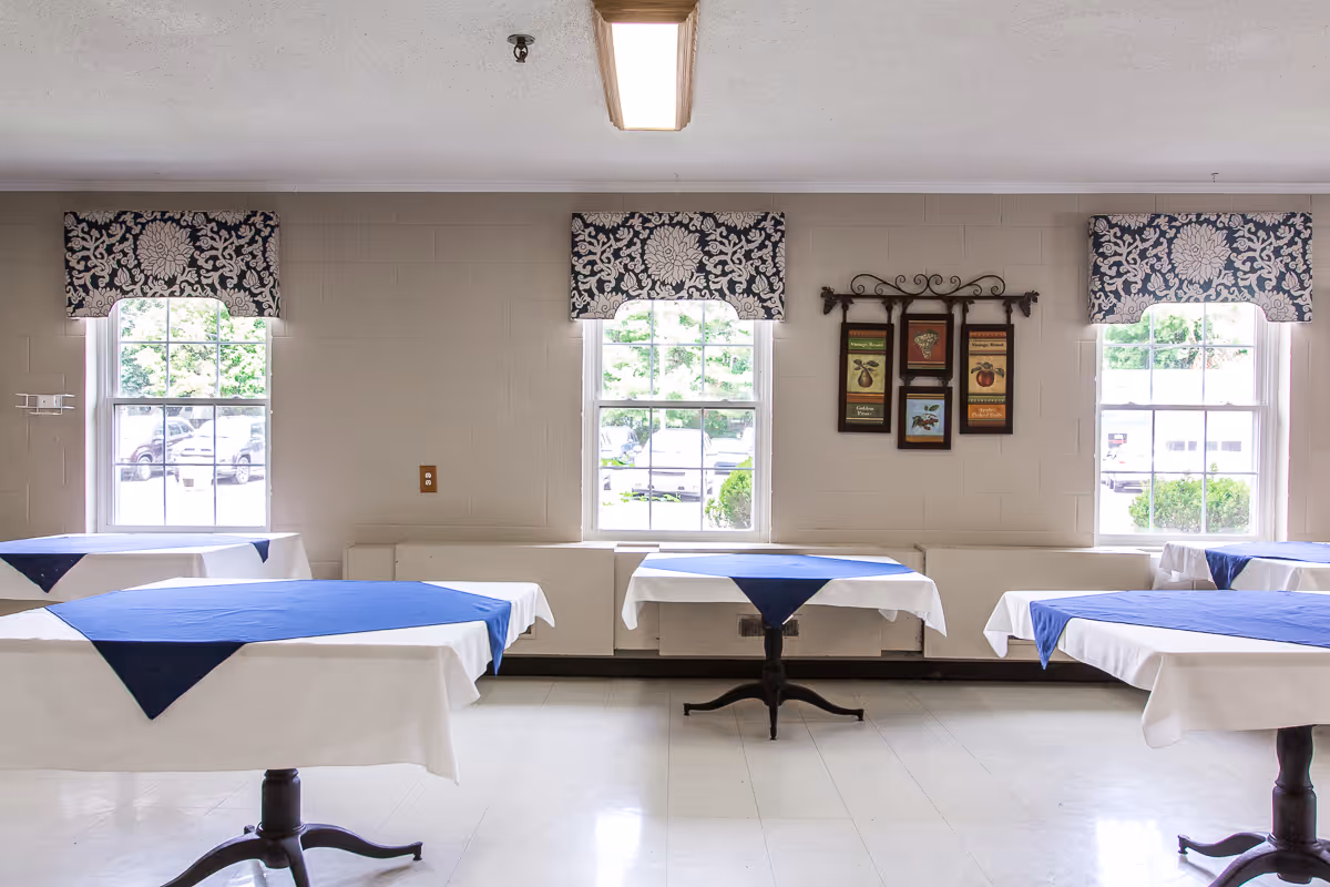 A bright dining room with several tables covered in white and blue tablecloths arranged in front of three windows.
