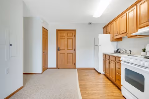Interior view of a senior living facility kitchen area with wooden cabinets, a white refrigerator, a white stove, and a sink. The floor transitions from wood in the kitchen area to carpet near the entrance door, which is wooden with a security lock. The walls are white and there is a fluorescent light fixture on the ceiling.