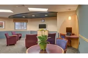 A cozy common area in a senior living facility with several maroon armchairs arranged around small tables. A round table with a green potted plant is in the foreground. There is a wall-mounted TV, a clock above it, and a small computer station in a curved alcove on the right side. The walls are painted in soft green and beige tones, and the carpet has a subtle pattern.
