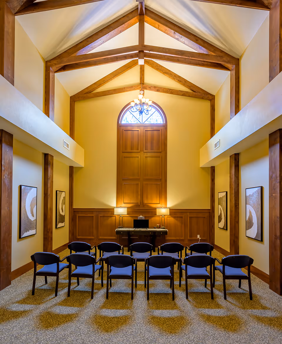 A well-lit room with a high vaulted ceiling featuring exposed wooden beams. The room has a large wooden panel on the far wall with an arched window above it. Below the window is a table with a laptop and two lamps. In front of the table, there are two rows of blue cushioned chairs arranged facing the table. The walls are decorated with framed abstract artwork on both sides.