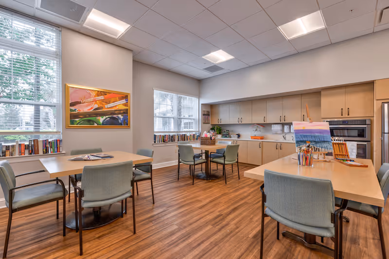 A bright and spacious activity room with wooden flooring, several tables and chairs, large windows letting in natural light, and a kitchen area with cabinets, a microwave, and a refrigerator. One table has art supplies and a painting on an easel, while books line the windowsills.