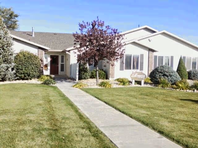 Single-story building with a gray roof and white exterior walls, surrounded by green grass, shrubs, and a tree with purple leaves. A concrete walkway leads to the entrance door. A sign near the entrance reads 'Rosetta'.