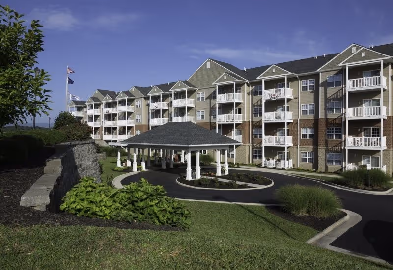 Exterior view of a multi-story senior living facility with beige and brick facade, multiple balconies, a covered circular driveway entrance, landscaped greenery, and flags flying on flagpoles under a clear blue sky.