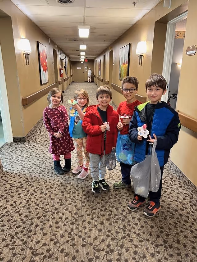 Five children standing in a carpeted hallway of a facility, smiling and holding small items. The hallway has beige walls with framed artwork and wall-mounted lights. A person is visible in the background walking away.