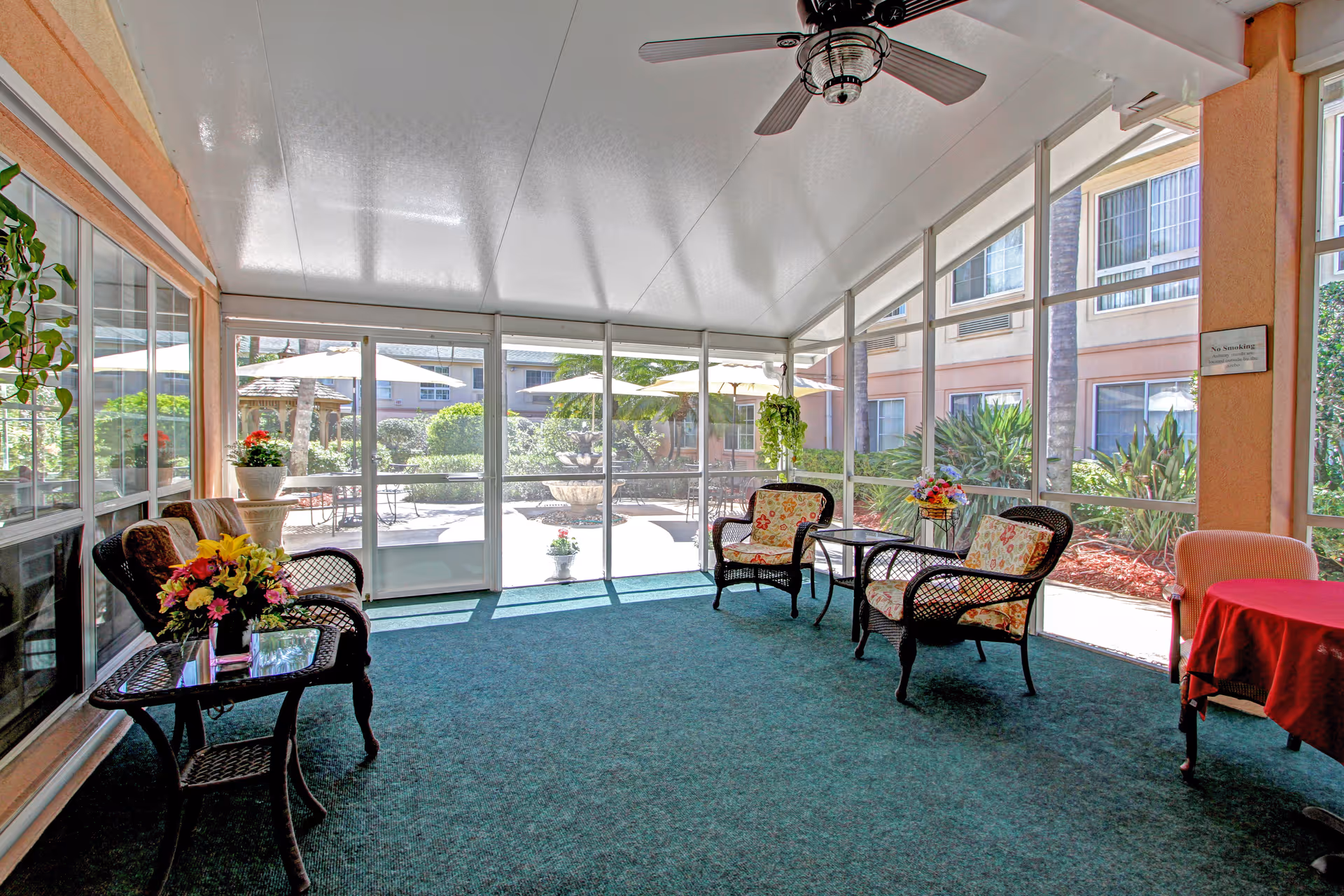 Sunroom with wicker chairs and tables, a ceiling fan, and large screened windows overlooking a courtyard with umbrellas and plants.
