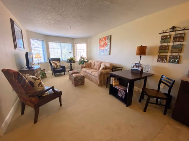 A cozy living room with beige carpet and cream-colored walls. The room features a striped sofa with a matching ottoman, a dark wooden armchair with floral cushions, and a black armchair near a window with white blinds. There is a small dark wooden table with a lamp and decorative items, a TV on a stand, and wall art including a colorful painting and a decorative hanging piece.