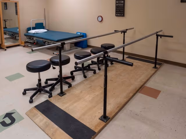 Physical therapy room with parallel bars for walking exercises, four black rolling stools, a padded examination table with a pillow, a mirror on the wall, a clock, and a motivational sign on the beige wall.