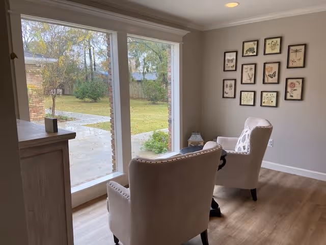 Two upholstered armchairs face a large window overlooking a backyard, with framed botanical prints on the wall.