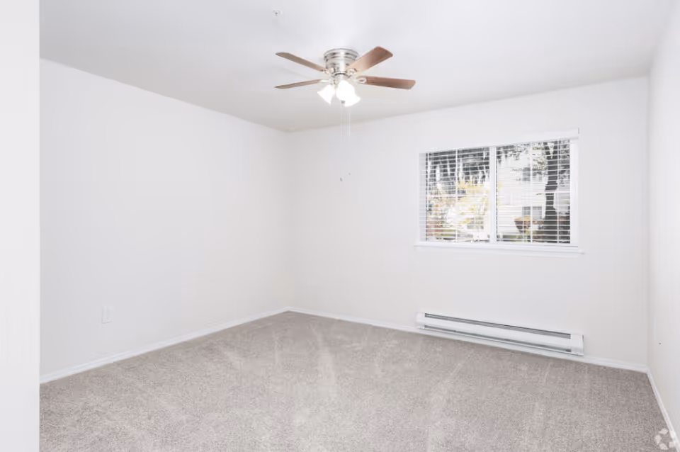 Empty bedroom with beige carpet, white walls, a ceiling fan with lights, and a window with white blinds showing trees outside.