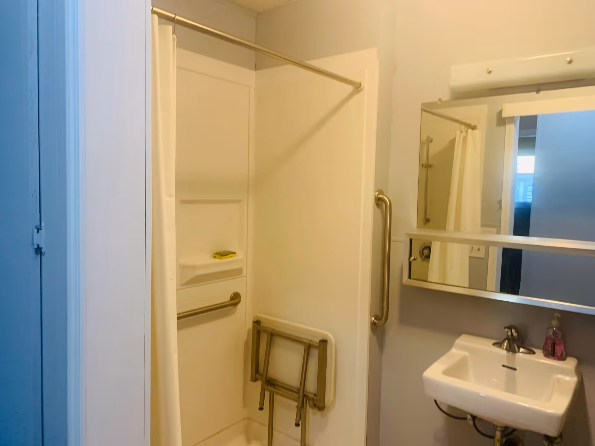 A small bathroom with a white shower area featuring a foldable shower seat and grab bars for accessibility. There is a white shower curtain on a rod, a small white sink with a faucet, a soap dispenser, and a mirrored medicine cabinet above the sink. The walls are painted light gray.