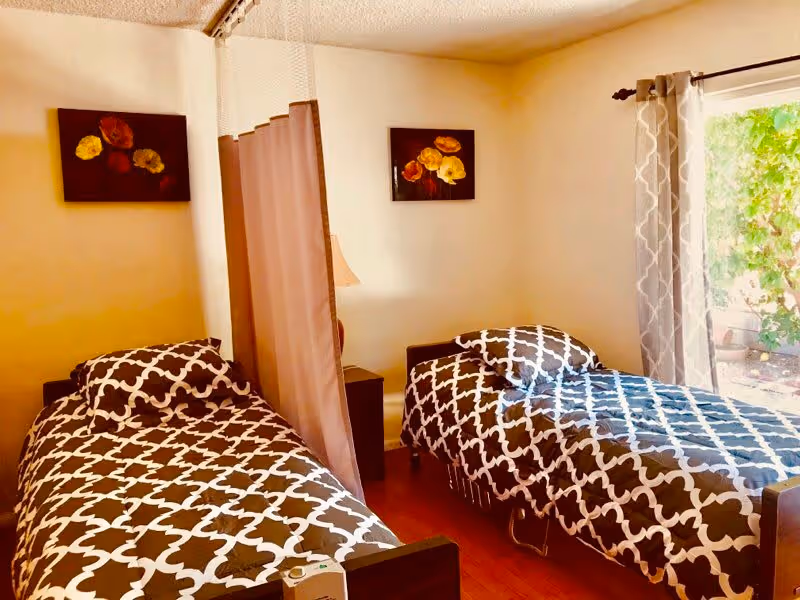 A room with two single beds separated by a privacy curtain. Each bed has a black and white patterned bedspread and pillow. There is a small nightstand with a lamp between the beds. Two framed floral paintings hang on the wall above the beds. A window with patterned curtains shows greenery outside.