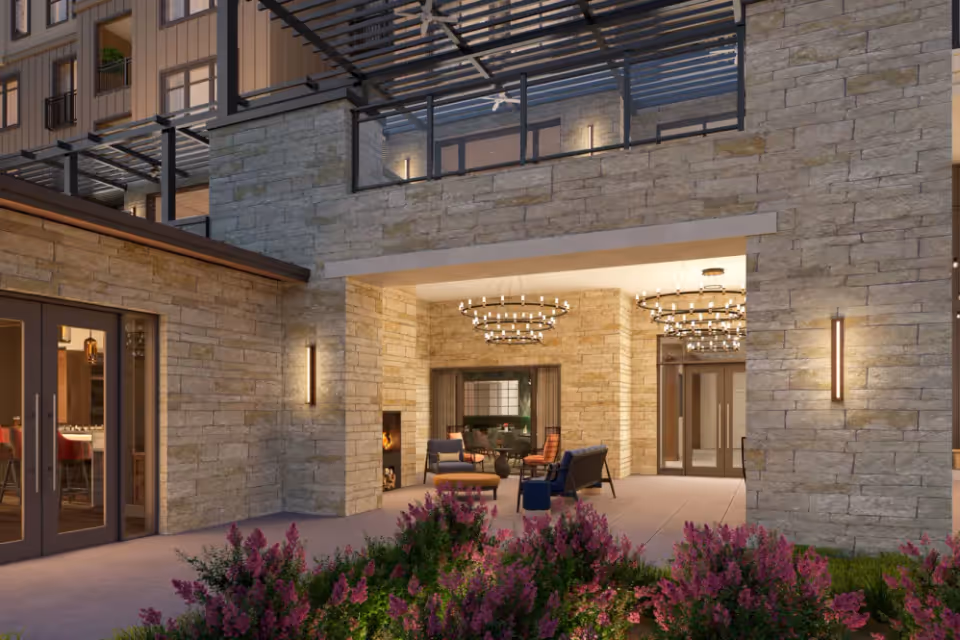 Stone-clad covered entry and outdoor lounge with chandeliers, seating, and flowering shrubs in front.