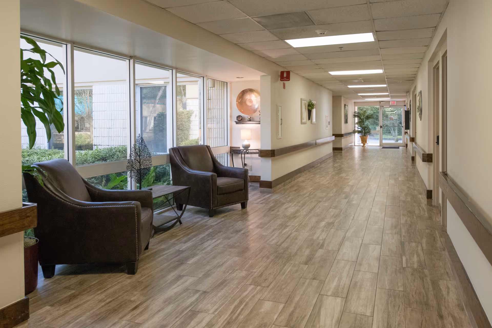Well-lit interior hallway with two armchairs by large windows, decorative plants and wood-look tile flooring.