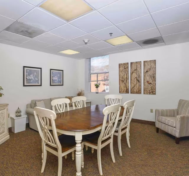 A cozy interior room with a wooden dining table surrounded by six white chairs. There is a beige sofa against the wall with two framed floral pictures above it. A window with blinds lets in natural light, and three vertical paintings of birds on branches hang on the adjacent wall. A striped armchair is positioned near the wall, and the room has a carpeted floor and a drop ceiling with fluorescent lights.