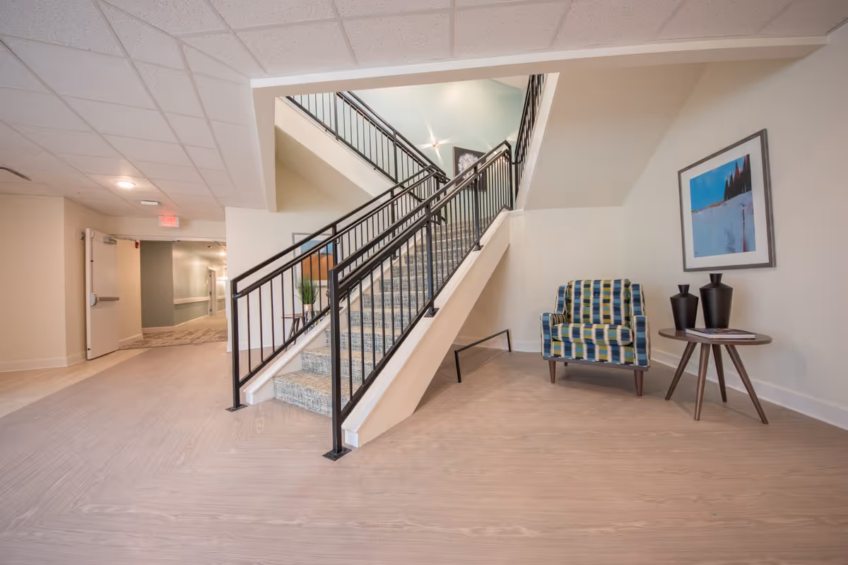Interior common area with a central staircase, a patterned armchair and side table under framed artwork and a hallway to the left.