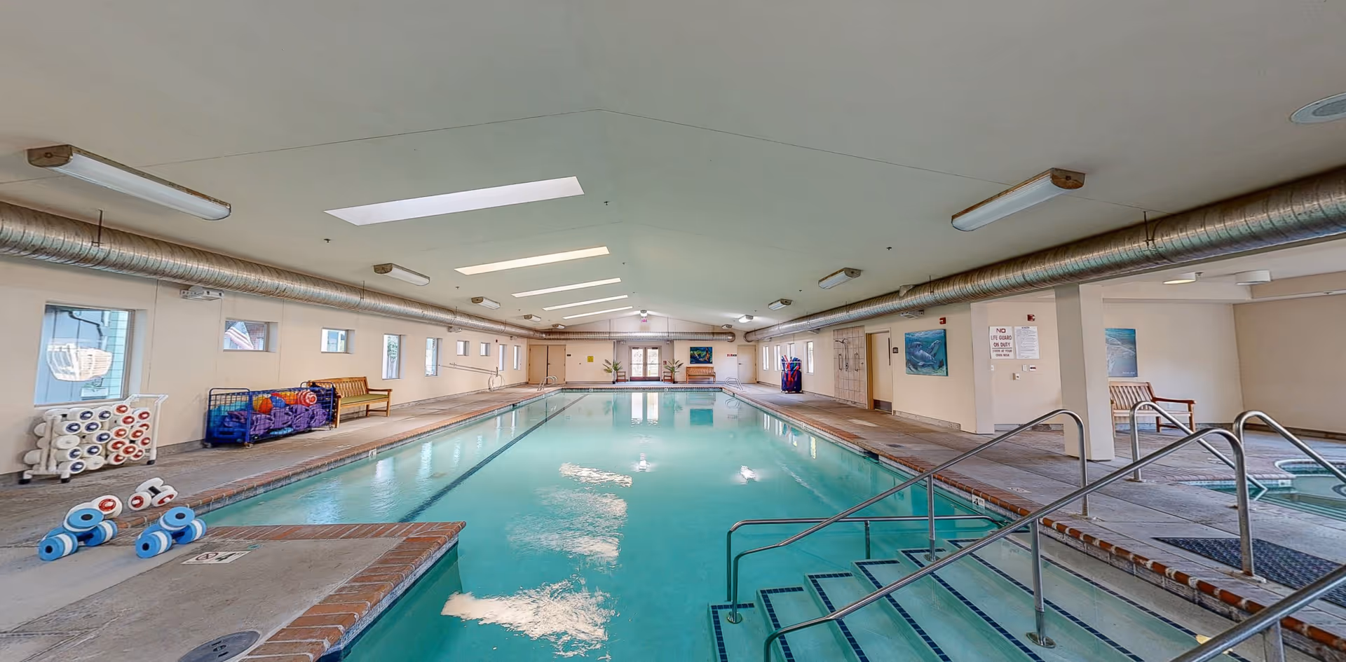 Indoor swimming pool with clear water, metal handrails, and steps leading into the pool. The pool area has a high ceiling with skylights and fluorescent lights. There are benches along the walls, pool equipment such as foam dumbbells and balls stored in racks, and several windows letting in natural light. A hot tub is visible to the right side of the pool.
