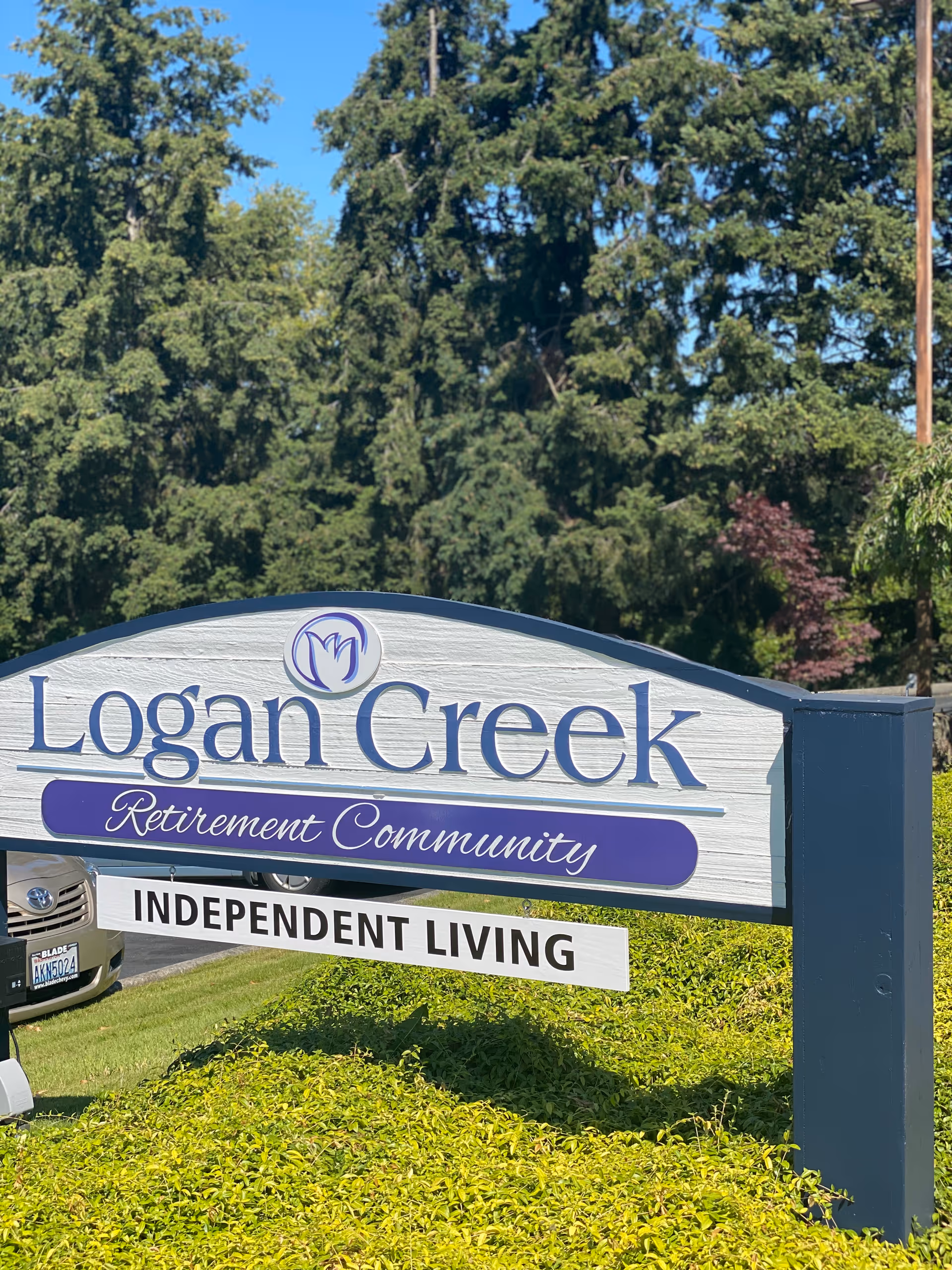 Outdoor sign for Logan Creek Retirement Community with the words 'Independent Living' below it, surrounded by greenery and trees in the background under a clear blue sky.