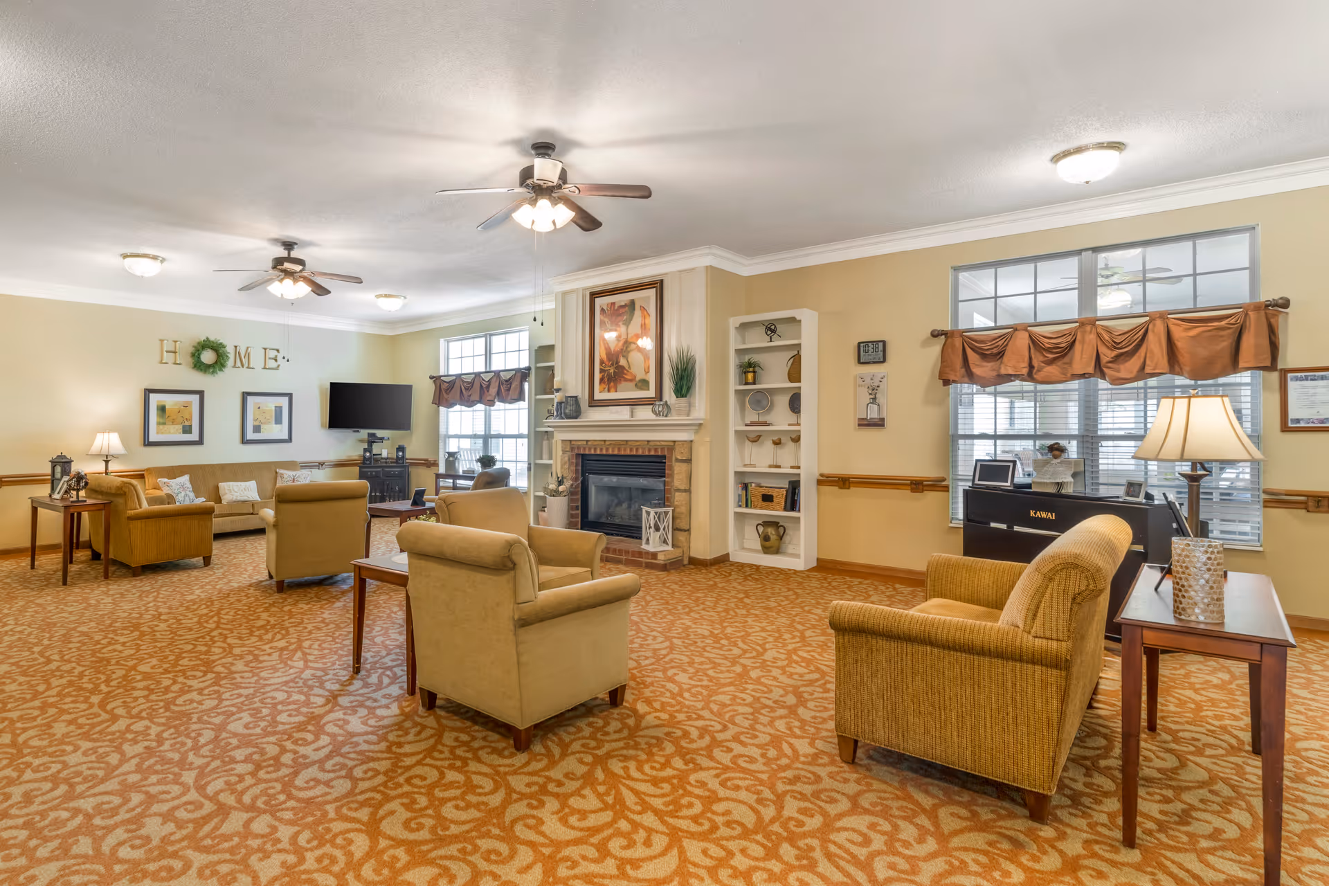 A spacious living room area with beige walls and patterned carpet. The room features multiple armchairs and sofas arranged around a central fireplace with a framed artwork above it. There are ceiling fans with lights, large windows with brown valances, a piano, and various decorative items on shelves and tables. The word 'HOME' is displayed on the wall above two framed pictures.