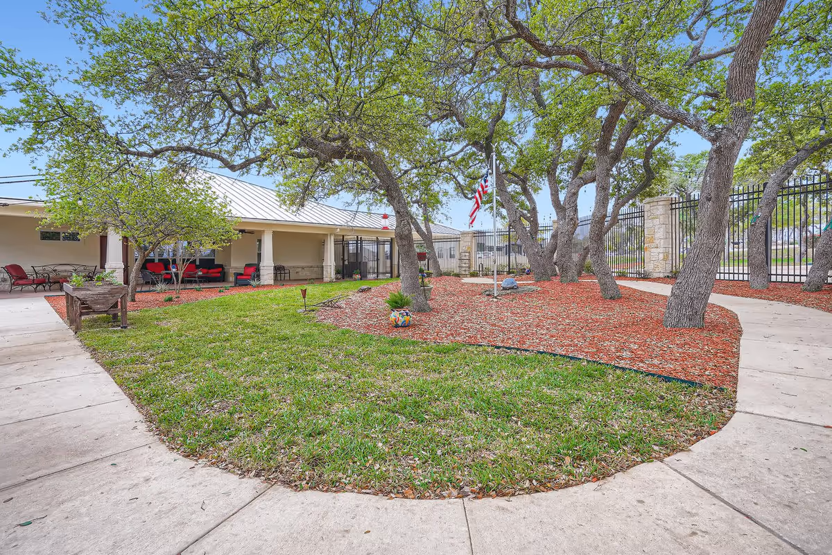 Outdoor courtyard area at OneStone Oak Senior Living featuring a curved concrete walkway surrounding a grassy area with several mature trees and mulch beds. There is a covered patio with red cushioned seating and an American flag displayed near a black metal fence.