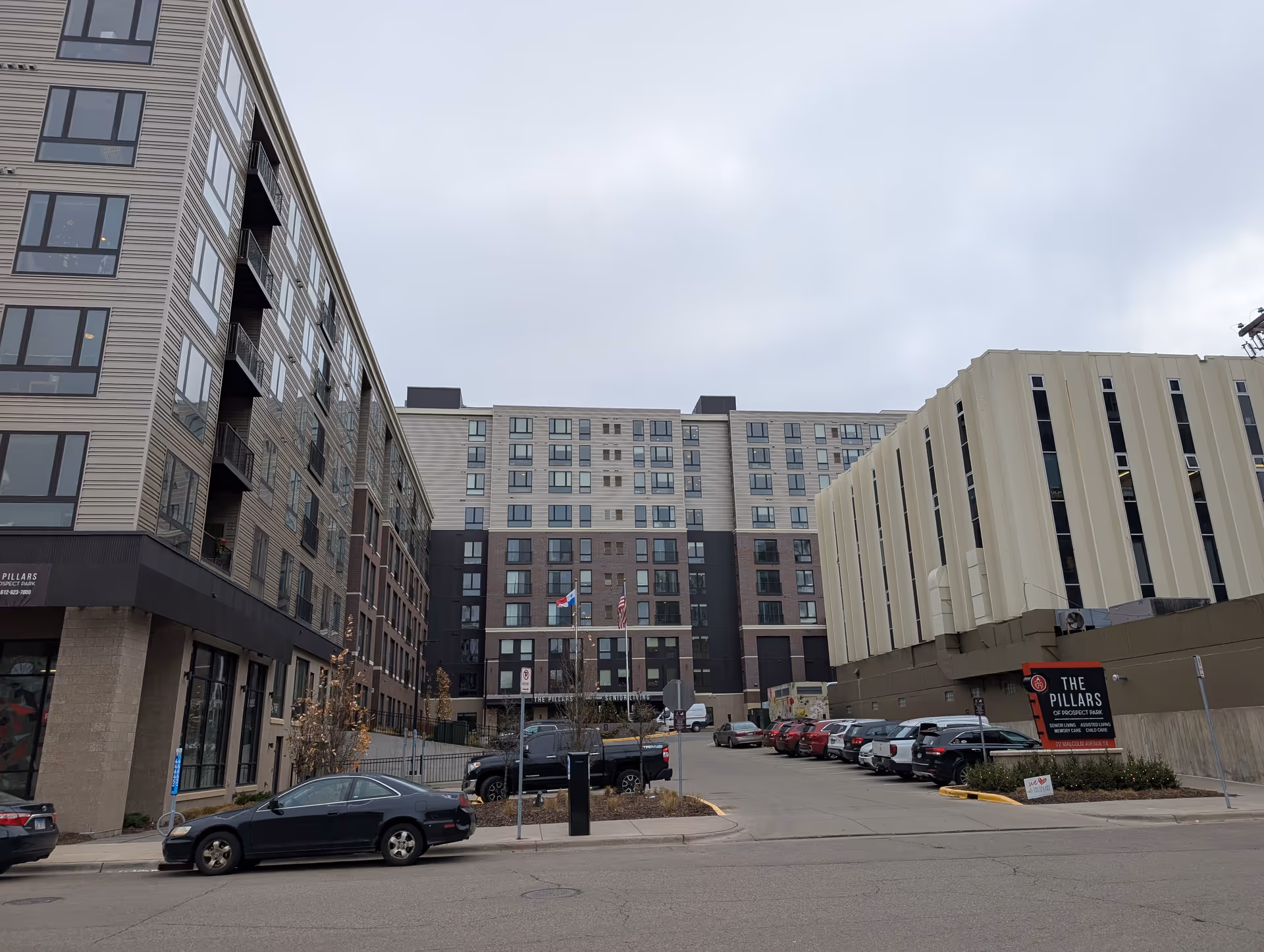 Exterior street view of a multi-story senior living building with parked cars and a sign reading 'The Pillars'.