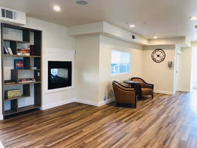 Bright communal lounge with wood-look floors, a wall-mounted fireplace, bookshelf, two armchairs with a small table, and a wall clock.