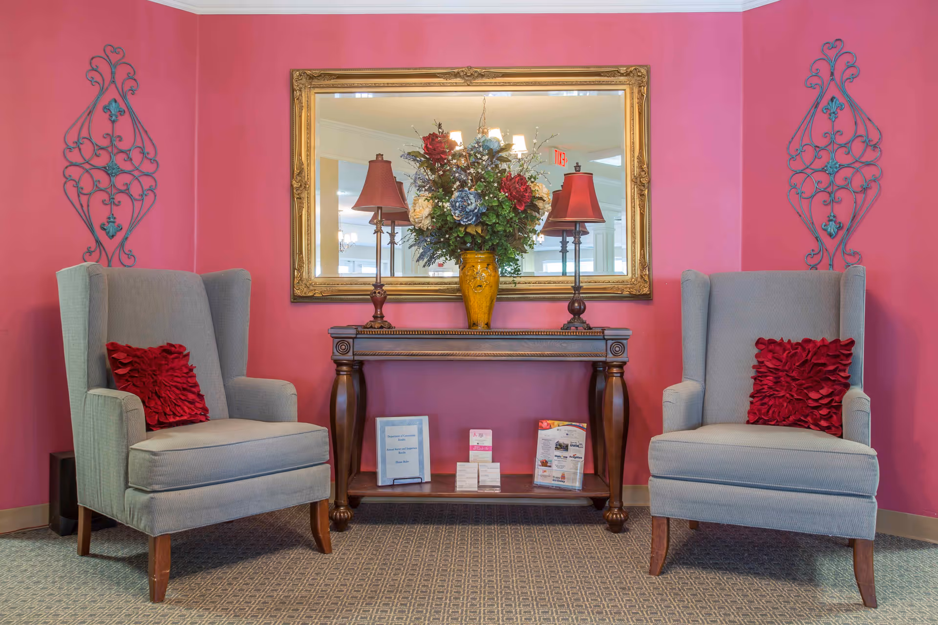 Two upholstered armchairs flank a wooden console table topped with a large floral arrangement and mirror against a pink wall.