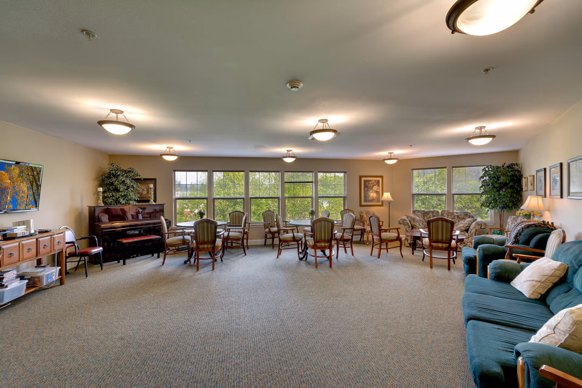 A spacious common area in a senior living facility with multiple chairs and small tables arranged near large windows that let in natural light. The room features a piano, a television mounted on the wall, several framed pictures, potted plants, and comfortable sofas with cushions. The ceiling has multiple light fixtures providing warm lighting.