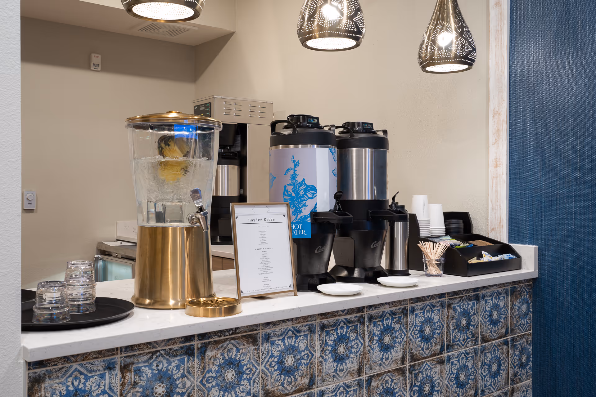 A beverage station with a large glass water dispenser containing lemon slices, two large coffee dispensers, a coffee maker, disposable cups, stirrers, and a tray with sugar and cream packets on a countertop with blue patterned tiles. Three decorative pendant lights hang above the station.