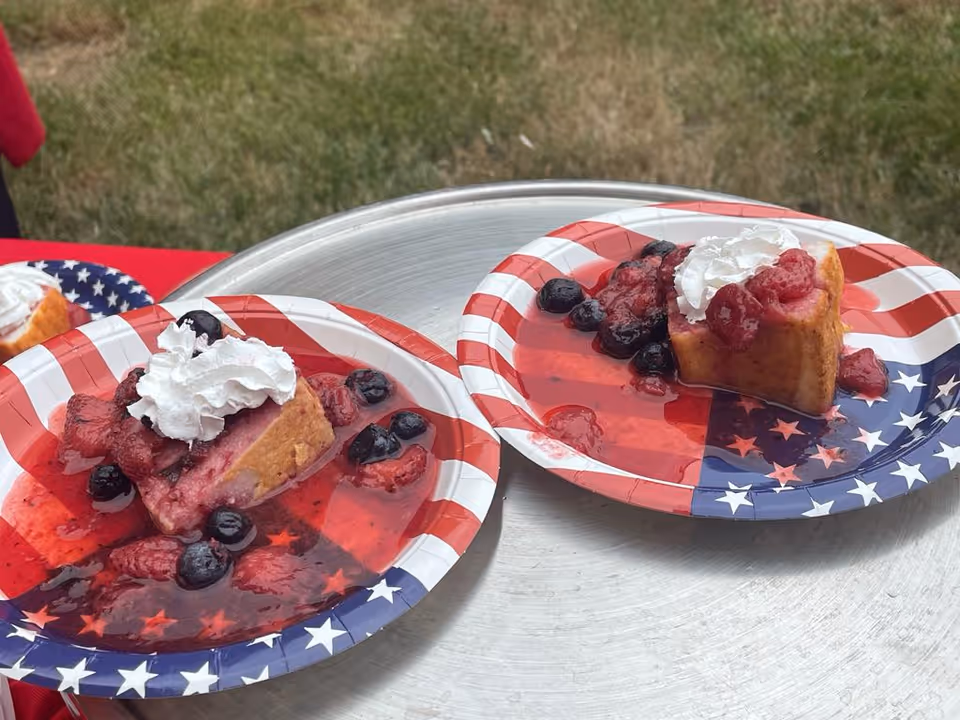 Two paper plates with an American flag design each holding a slice of cake topped with whipped cream and mixed berry sauce, placed on a round metal tray outdoors with grass in the background.